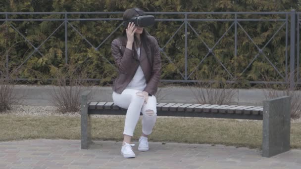 mujer joven que obtiene experiencia en el uso de auriculares VR o auriculares de realidad virtual al aire libre mientras está sentada en el banco en el parque
 - Imágenes, Vídeo