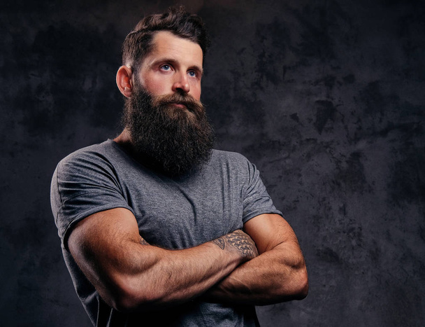 Portrait en gros plan d'un hipster à la barbe pleine et à la coupe de cheveux élégante, vêtu d'un t-shirt gris, debout avec les bras croisés dans un studio sur un fond sombre
. - Photo, image