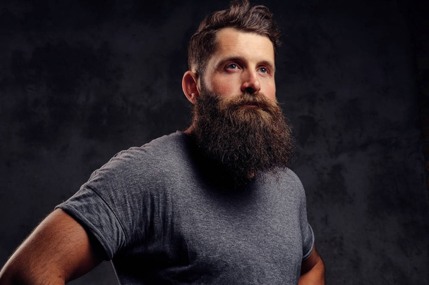 Portrait en gros plan d'un hipster à la barbe pleine et à la coupe de cheveux élégante, vêtu d'un t-shirt gris, debout dans un studio sur un fond sombre
. - Photo, image