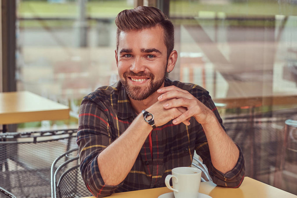 A joyful handsome fashionable male with stylish haircut and beard, wearing fleece shirt, drinking coffee at the cafe. - Foto, afbeelding