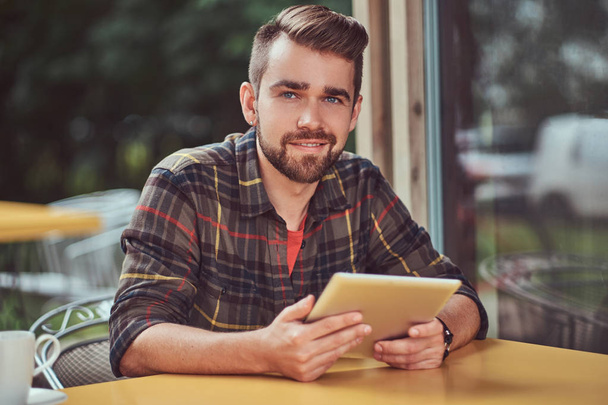 Un guapo y sonriente freelancer masculino de moda con elegante corte de pelo y barba, con camisa polar, trabajando en una tableta dentro de una cafetería
. - Foto, imagen