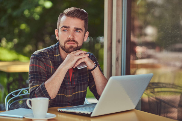 Een lachende knappe modieuze mannelijke freelancer met stijlvolle kapsel en baard, het dragen van fleece shirt, werken op een laptopcomputer en een smartphone binnen een cafetaria te houden. - Foto, afbeelding