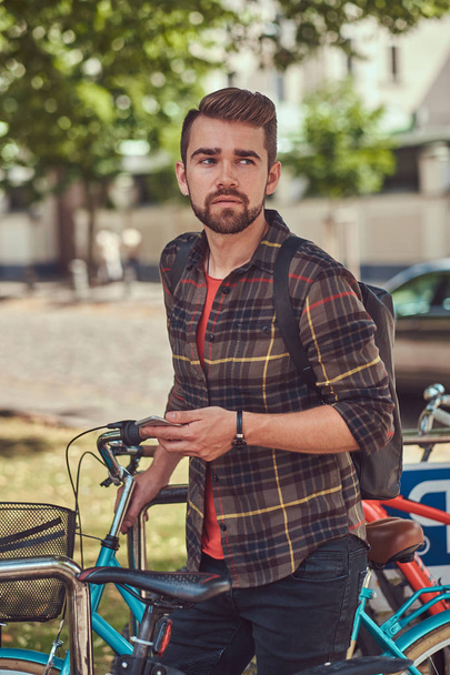A handsome fashionable young male with stylish haircut and beard, wearing fleece shirt, holds a smartphone,  stands in the parking lot for bicycles in the park. - Fotografie, Obrázek