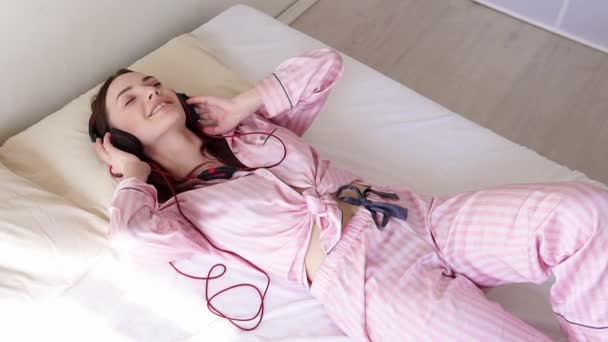 woman in pajamas is lying on the bed dances to music from headphones - Materiał filmowy, wideo