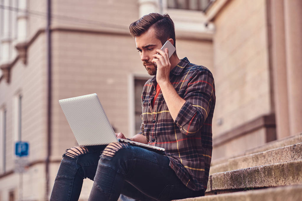 A beautiful freelancer with stylish haircut and beard, wearing a fleece shirt and jeans, working on a laptop computer and talk by a smartphone, sitting on steps against an old building. - Photo, Image