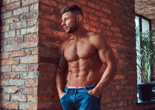 Porträt eines kräftigen, gutaussehenden, hemdlosen Mannes mit muskulösem Körper, der Jeans trägt und sich in einem Raum mit Dachboden an eine Ziegelwand lehnt. - Foto, Bild