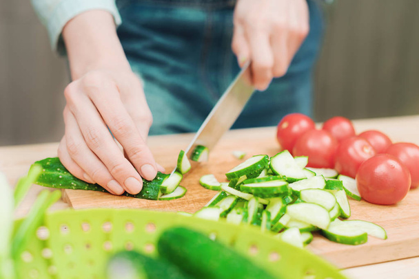 close-up of female hands cut into fresh cut cucumbers on a wooden cutting board next to pink tomatoes. The concept of homemade vegetarian cuisine and healthy eating and lifestyle - Photo, image