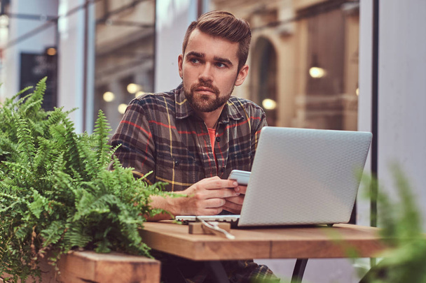 Portrait of a young freelancer with stylish haircut and beard, dressed in a fleece shirt, using a smartphone while working on a laptop computer, sitting in a cafe outdoors. - Photo, Image