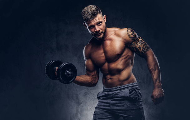 Powerful stylish bodybuilder with tattoo on his arm, doing the exercises on a biceps with dumbbell, look at the camera with a confident look. Isolated on a dark background. - Foto, Imagen