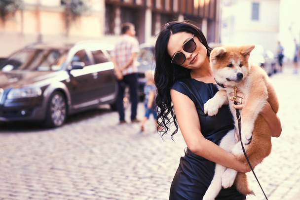Стильная брюнетка в черном платье и солнечных очках, стоящая на улице и держит милого чистокровного щенка Акита
. - Фото, изображение