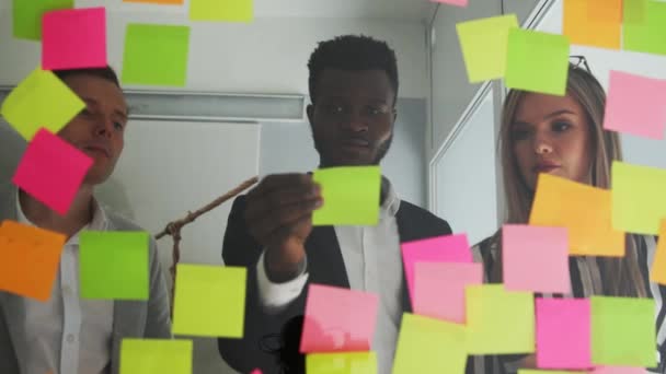 Multi ethnic creative team conducts brainstorming all their ideas are pasted on colored stickers on the glass wall. African American male and blonde team up in the office - Footage, Video