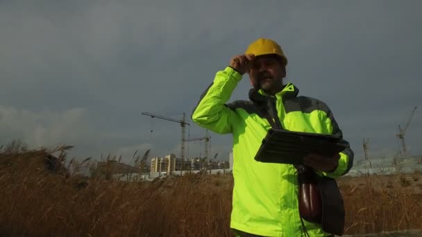 dojrzały mężczyzna robotnik majster w hełm z tabletem na tle budowy - Materiał filmowy, wideo