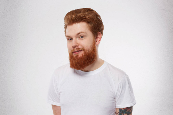 Young redhead man with heavy, massive beard smiles and feels happy. Portrait of pleased, joyful male. Isolated over studio white wall - Photo, Image