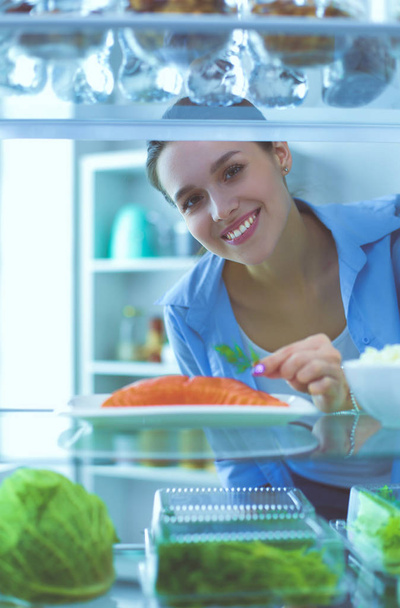 Portrait of female standing near open fridge full of healthy food, vegetables and fruits - 写真・画像