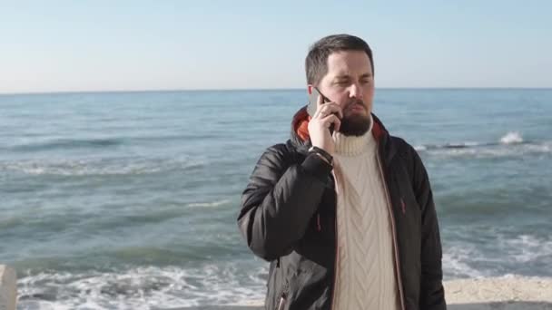 взрослый мужчина, одетый в осеннюю куртку, разговаривает по мобильному телефону у моря
 - Кадры, видео