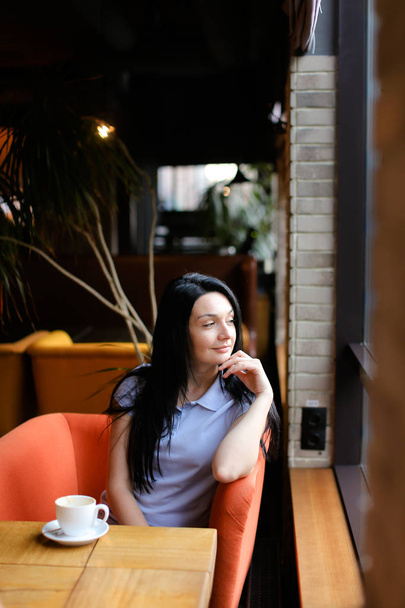 Thinking woman sitting in orange arm chair at cafe with cup of coffee. - Fotó, kép