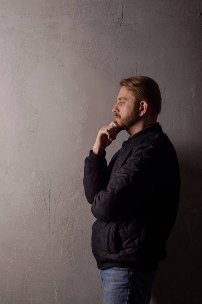 Бородатий чоловік середнього віку в чорній куртці і джинсах стоїть біля стіни, дивлячись далеко
 - Фото, зображення