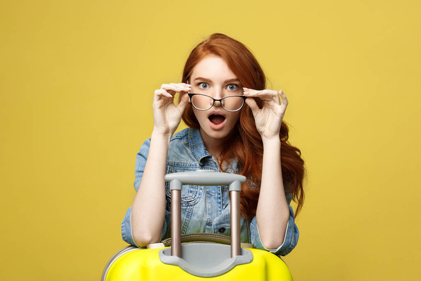 Travel and Lifestyle Concept: Portrait of a shocked girl in denim dress with suitcase looking at camera isolated over golden yellow background - Foto, Imagem
