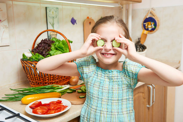 Girl closes her eyes with cucumbers and have fun in home kitchen. Basket of vegetables and fresh fruits in kitchen interior. Healthy food concept - Fotoğraf, Görsel