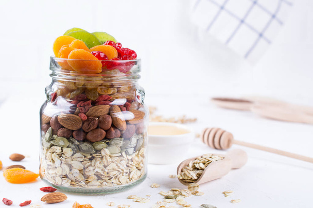 Glass jar with ingredients for cooking granola on white background. Oat flakes, honey, nuts, dried fruit and seeds. Healthy snak - Fotografie, Obrázek