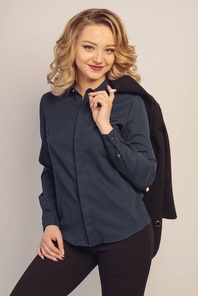 Портрет улыбающейся красивой молодой деловой женщины, стоящей с ручной курткой за плечом на сером фоне и позирующей модной
. - Фото, изображение