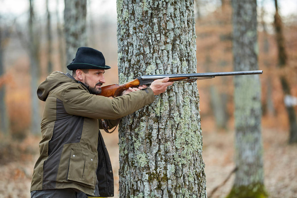 Hunter with double barrel gun hiding behind tree and aiming at wild animal - Фото, зображення