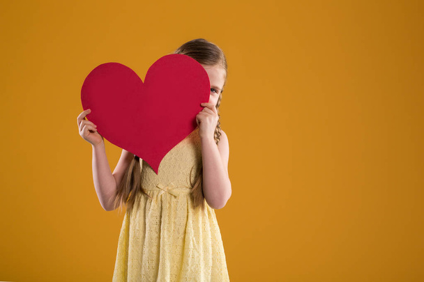 Young girl in yellow dress peeking behind red heart - Foto, imagen
