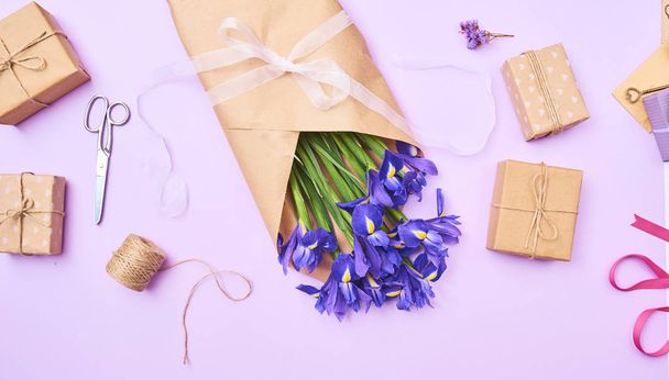 Поздравительная открытка с весенне-фиолетовыми цветами радужной оболочки и крошечной подарочной коробочкой, ножницами, лентой на фиолетовом фоне, местом для текста. вид сверху
 - Фото, изображение
