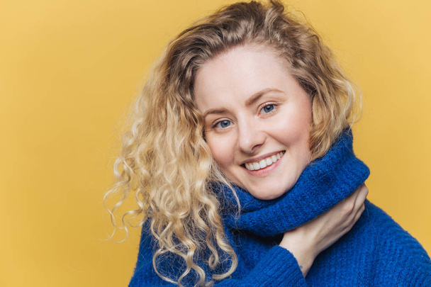 Close up shot of delighted satisfied female model has curly blonde hair, broad smile, demonstrates white perfect teeth, wears blue knitted sweater, isolated over yellow background. Happiness - 写真・画像