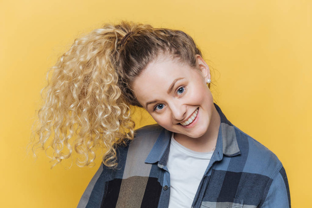 Funny cute female model with curly blonde pony tail, has positive smile, has fun alone indoor against yellow background, wears casual checkered shirt, being in good mood. Facial expressions. - Photo, Image
