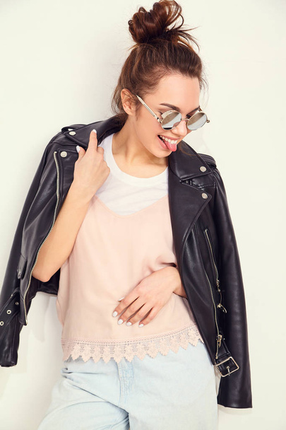 Portrait of young beautiful brunette woman girl model with nude makeup wearing summer hipster biker leather jacket clothes in sunglasses posing near wall. Going crazy and showing her tongue - Foto, immagini
