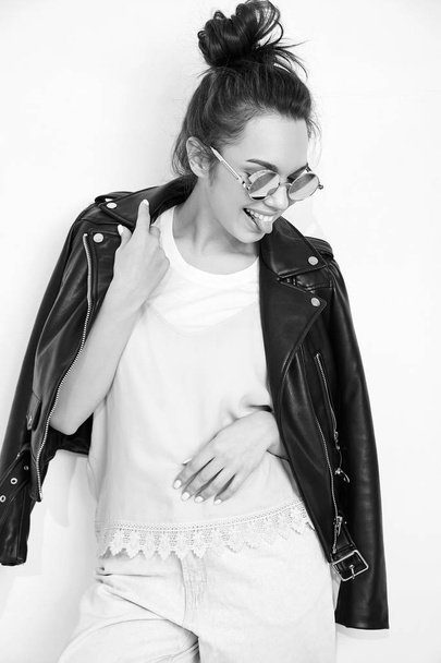 Portrait of young beautiful brunette woman girl model with nude makeup wearing summer hipster biker leather jacket clothes in sunglasses posing near wall. Going crazy and showing her tongue - 写真・画像