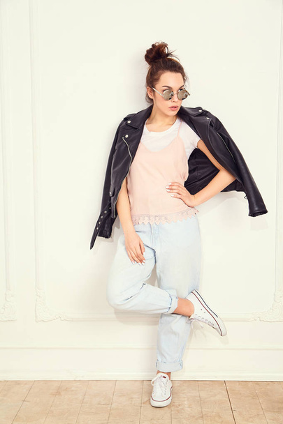 Portrait of young beautiful brunette woman girl model with nude makeup wearing summer hipster biker leather jacket clothes in sunglasses posing near wall. Going crazy  - Foto, immagini