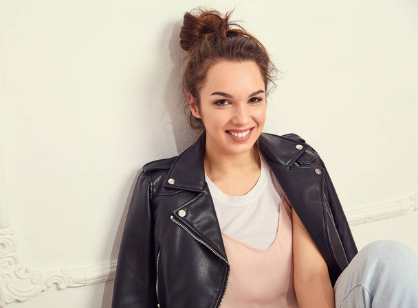 Portrait of young beautiful brunette woman girl model with nude makeup wearing summer hipster biker leather jacket clothes posing near wall. Sitting on the floor - Foto, Imagem