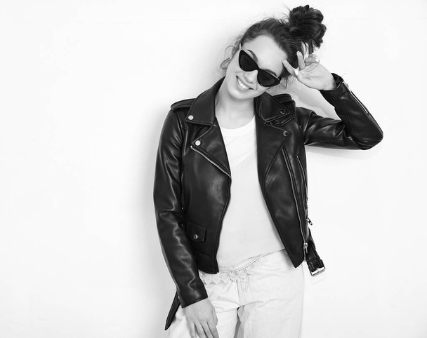 Portrait of young beautiful brunette woman girl model with nude makeup wearing summer hipster biker leather jacket clothes in sunglasses posing near wall. Going crazy  - Photo, image