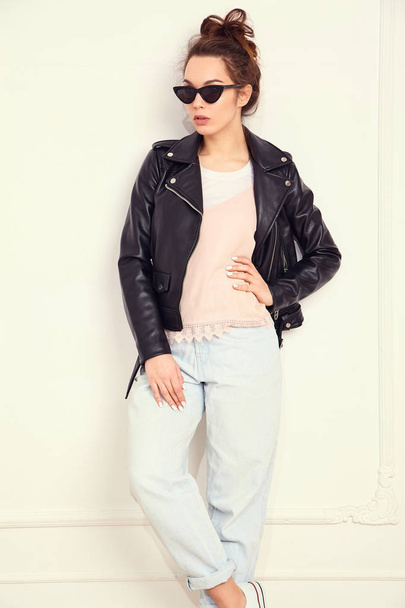 Portrait of young beautiful brunette woman girl model with nude makeup wearing summer hipster biker leather jacket clothes in sunglasses posing near wall. Going crazy  - Foto, imagen