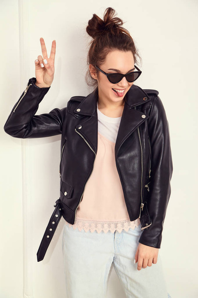 Portrait of young beautiful brunette woman girl model with nude makeup wearing summer hipster biker leather jacket clothes in sunglasses posing near wall. Showing her tongue and peace sign - Valokuva, kuva