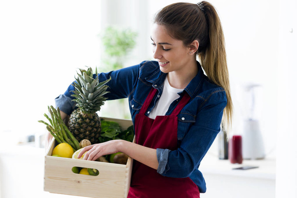Pretty young woman holding a box with fruits and vegetables in the home kitchen. - Photo, Image