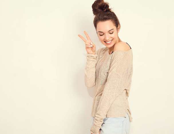 Portrait of young beautiful brunette woman girl model with nude makeup in summer hipster clothes posing near wall. Going crazy and showing peace sign - 写真・画像