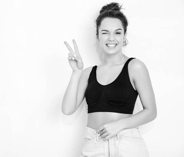 Portrait of young beautiful brunette woman girl model with nude makeup in summer T-shirt top and jeans hipster clothes posing near wall. Winking and showing peace sign - 写真・画像