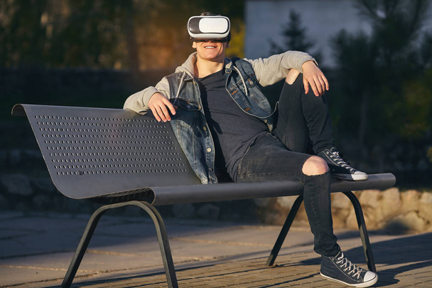 Blurred lines of reality.  The young men sits on the bench and wears VR glasses and looking out and smiling. Outdoor. Copy space. - Foto, Imagem