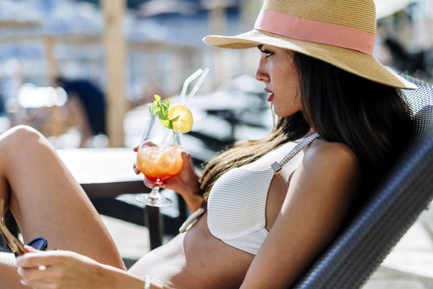 Stunning beauty drinking her cocktail and sunbathing while wearing a hat and a white bikini - Фото, изображение