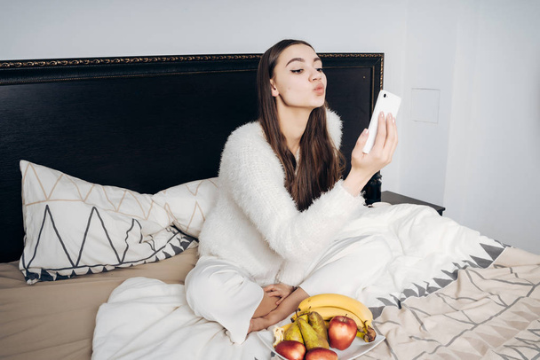cute young girl in pajamas resting in bed after working day, making selfie on her smartphone - Фото, зображення