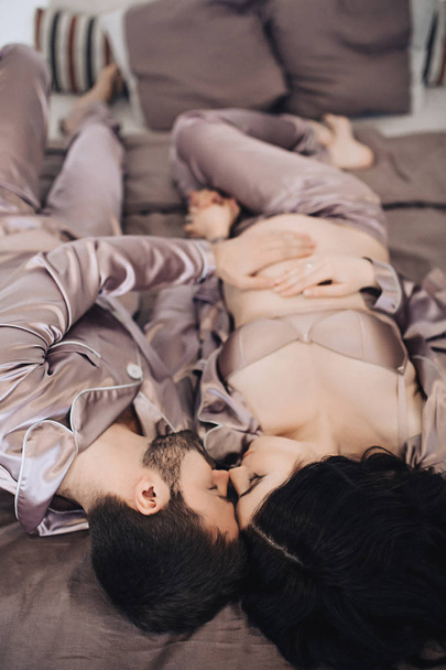 Happy future parents lay on the bed and hug in pajamas, parents are waiting for baby, married couple is waiting for a child, pregnant woman with long hair, beauty of pregnancy, love and support in - Photo, Image