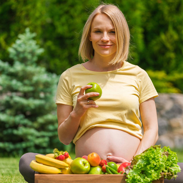 Pregnant woman sitting on grass and holding an apple in hand - Fotoğraf, Görsel