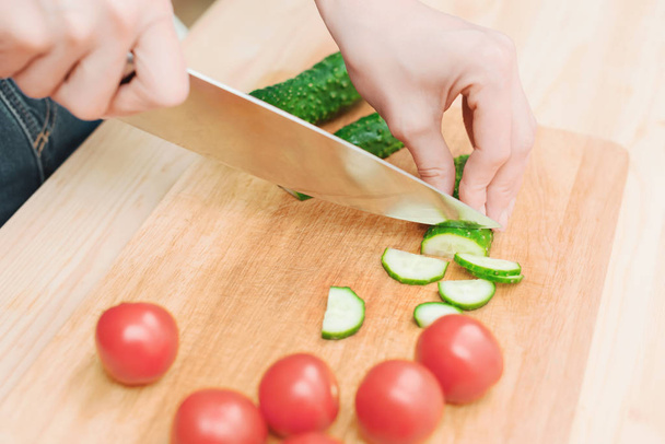 close-up of female hands cut into fresh cut cucumbers on a wooden cutting board next to pink tomatoes. The concept of homemade vegetarian cuisine and healthy eating and lifestyle - Foto, afbeelding