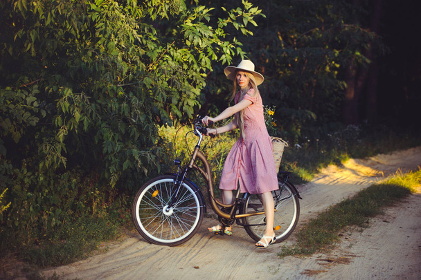 Belle fille portant une jolie robe rose s'amusant dans un parc avec un vélo tenant un beau panier avec des fleurs. Paysages anciens. Jolie blonde au look rétro, vélo et panier à fleurs
 - Photo, image