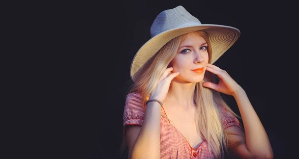Beautiful young woman in pink dotted summer dress and sun hat points to copy space, looking at camera. Three quarters of a studio length shot on a dark natural background - Photo, Image
