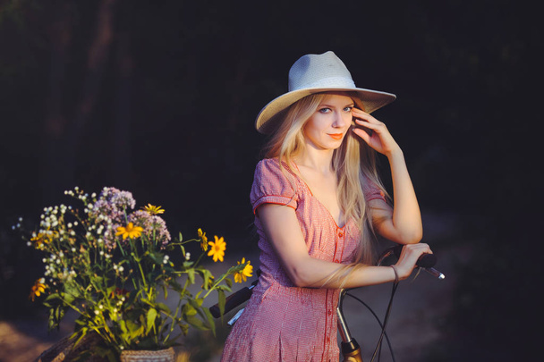 Beautiful girl wearing a nice pink dress having fun in a park with a bicycle holding a beautiful basket with flowers. Vintage landscapes. Pretty blonde with retro look, bicycle and basket with flowers - Foto, imagen