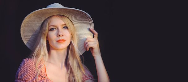 ピンクの点線の夏のドレスと太陽の帽子の美しい若い女性はカメラを見て空間をコピーを指します。暗い自然背景に撮影スタジオの長さの 4 分の 3 - 写真・画像